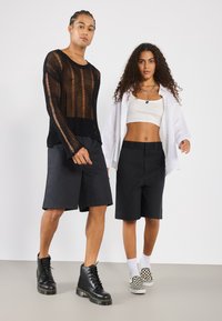 Haut en maille noire transparente à manches longues, short noir, bottes de combat noires, crop top blanc, chemise blanche surdimensionnée, tennis à carreaux noir et blanc.