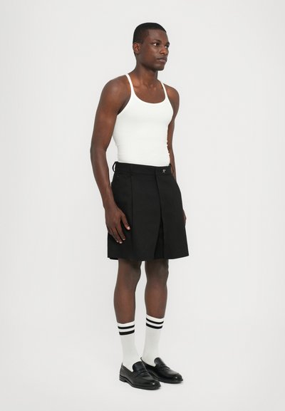 Schwarze plissierte Shorts mit seitlichen Taschen und Knopfverschluss, kombiniert mit einem weißen Tanktop, schwarzen Loafers und weißen Socken mit schwarzen Streifen.