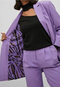 Blazer lavande avec une doublure intérieure noire présentant un motif abstrait violet et marron, assorti à un haut noir et un pantalon lavande coordonné.