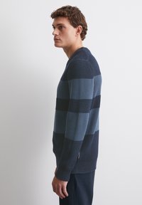 Herren-Pullover mit langen Ärmeln, der strukturierte Streifen in Dunkelblau und Hellblau aufweist. V-Ausschnitt-Design mit seitlichen Schlitzdetails. Gewebte Stoffstruktur.