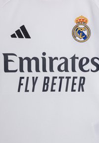 Camiseta deportiva blanca con el texto en negro "Emirates Fly Better". Logotipo de Adidas en negro a la izquierda; emblema bordado del Real Madrid con una corona a la derecha.