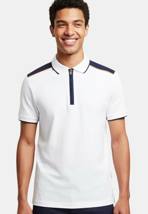 Homme souriant portant un polo blanc avec des rayures épaules bleu marine et orange et une courte fermeture éclair à l'avant, debout devant un fond uni.