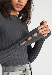 Haut gris à manches longues côtelées avec des détails découpés et des accents de clous noirs sur les manches. Associé à un pantalon à motif camouflage.