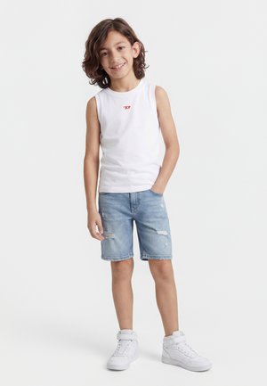 Dreng med brunt hår iført hvid ærmeløs skjorte, lyseblå revnede denimshorts og hvide højtop-sneakers, stående med den ene hånd i lommen.