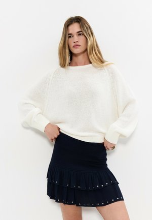 Femme portant un pull blanc surdimensionné et une mini-jupe à volants noire ornée de petits clous métalliques, debout devant un fond blanc.