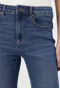 Jeans de denim azul com corte slim, apresentando fecho de correr, bolsos frontais e leve desbotamento no tecido. Detalhe de botão na cintura.