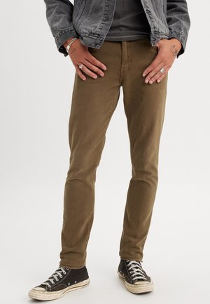 Person trägt braune Slim-Fit-Hose, schwarze High-Top-Sneaker, schwarzes T-Shirt und graue Jeansjacke, Hände ruhen auf den Hüften.