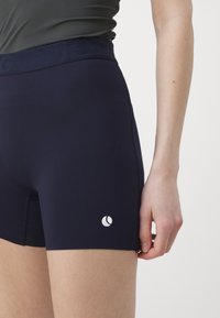 Ναυτικό μπλε αθλητικές βερμούδες με λεία υφή, διαθέτοντας φαρδύ λάστιχο και ένα μικρό λευκό λογότυπο στη χαμηλή αριστερή πλευρά.