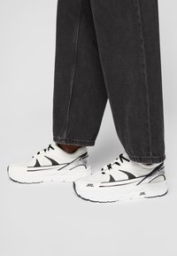 Hvite joggesko med svarte detaljer, teksturert mesh-overdel, avrundet tå, og tykk såle. Brukt med løse, mørke denimbukser.