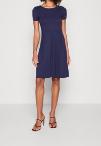 Vestito blu navy a maniche corte con corpino aderente e dettagli della gonna plissettata, realizzato in un tessuto morbido, abbinato a tacchi a strisce marroni.