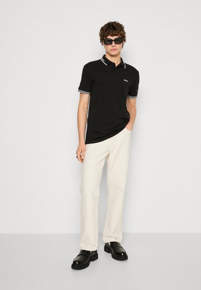 Polo noir avec des accents blancs, à manches courtes, et logo sur la poitrine ; associé à un pantalon beige clair et des chaussures noires.
