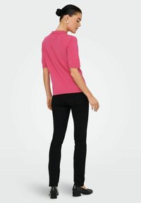 Donna con capelli scuri raccolti a chignon, che indossa un top a maglia rosa con maniche corte, pantaloni neri aderenti e scarpe nere con tacco basso, in piedi voltata di spalle.