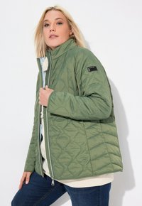 Giacca imbottita verde con colletto alto, zip frontale e tasche laterali. Presenta un'etichetta in tessuto sulla manica e una texture liscia.