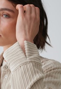 Beige, ribbed fabric with a textured finish. The collar features a wavy edge and a fitted cuff. Design prioritizes softness and comfort.