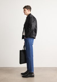 Chaqueta bomber negra, camisa blanca, jeans azules, botas negras, sosteniendo una bolsa tote negra. Diseño minimalista, textura suave, silueta ajustada.
