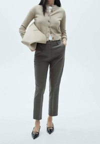 Gilet beige avec boutons, chemise blanche en dessous, pantalon gris ajusté et escarpins noirs à bout pointu. Portant un sac à main de couleur crème.