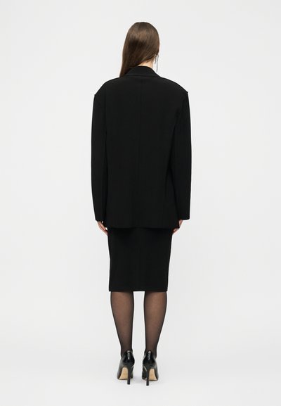 Γυναίκα που στέκεται με την πλάτη γυρισμένη, φοράει μαύρο υπερμεγέθες σακάκι, ταιριαστή pencil φούστα, διάφανα μαύρα καλσόν και μαύρες ψηλοτάκουνες γόβες.