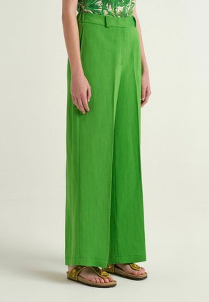 Donna che indossa pantaloni verde brillante a vita alta e gambe larghe, abbinati a sandali gialli con perline e una maglietta verde con motivo a fantasia.