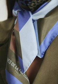 Écharpe en soie avec des motifs géométriques dans des tons de bleu, marron et vert, avec une texture lisse et un logo de marque visible en bas.