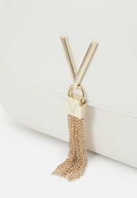 Bílá kabelka s akcentem ve tvaru V v zlatém tónu a zámkem s detailním řetízkovým třásněním. Hladký, strukturovaný materiál s elegantním designem.