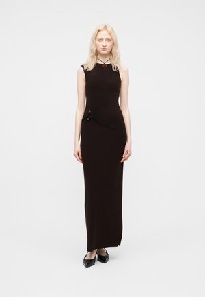 Blondynka stojąca, ubrana w czarną sukienkę maxi bez rękawów z marszczeniem po bokach i czarne spiczaste baleriny na białym tle.
