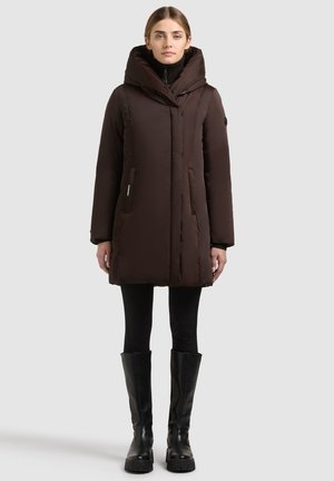 Femme portant un manteau matelassé marron foncé longueur genou avec un grand col, des leggings noirs et des bottes noires montantes, debout devant un fond uni.