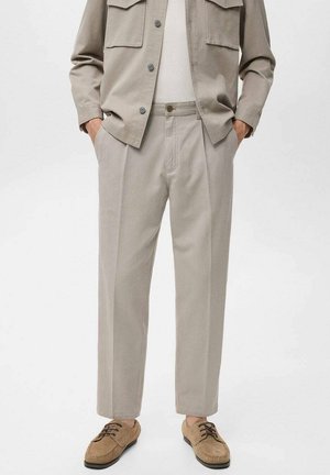 Beige, tailored pants with a straight cut, featuring front pleats and side pockets. Paired with a matching light jacket and classic shoes.