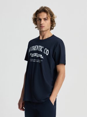 Młody mężczyzna o falowanych włosach, ubrany w granatowy T-shirt z białym napisem "Authentic Co Brooklyn NYC", stoi z jedną ręką w kieszeni na szarym tle.