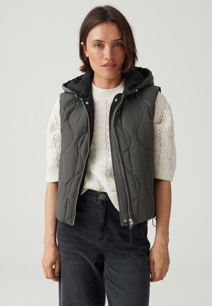 Woman wearing a dark gray quilted sleeveless hooded vest over a white knit sweater and black pants, standing against a plain background.