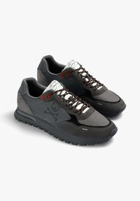 Zapatillas deportivas grises con material de malla y ante, que presentan una superficie texturizada, detalles en marrón y un logo de calavera. Los cordones marrones completan el diseño.