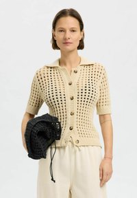 Cárdigan de punto beige de manga corta con un patrón estructurado, con botones en la parte delantera y un bolso texturizado negro en contraste sostenido en la mano.