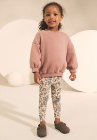 Roze sweatshirt met opgepofte mouwen, legging met bloemenpatroon in crème, blauw en bruin, en donkerbruine slip-on schoenen.