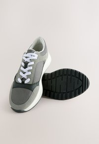 Grå og sort mesh-sneaker med hvide snørebånd og tyk hvid sål, en sko står oprejst, og den anden viser sort slidsolev.