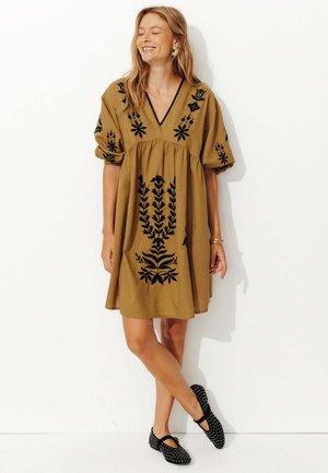 Robe brune mi-longue avec broderie florale noire, manches bouffantes et décolleté en V. Associée à des chaussures noires à enfiler avec des accents ronds.