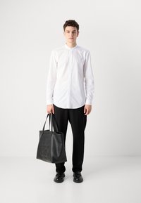 Joven con camisa blanca de botones y pantalones negros sosteniendo un gran bolso tote de piel negro con texto en relieve, de pie frente a un fondo blanco.
