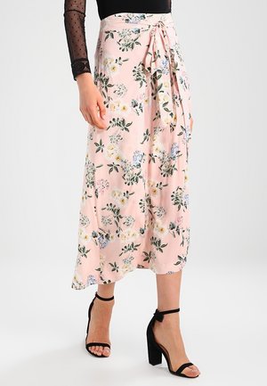 Kvinde iført en lyserød blomsterprintet midi-nederdel med bundet talje og sorte højhælede sandaler med ankelrem, kombineret med en gennemsigtig sort langærmet top.