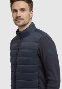 Marineblå puffervest med høj krave, vandret quiltning og ribstrikkede sweaterærmer. Har en sidelukning med lynlås og logo-patch på skulderen.