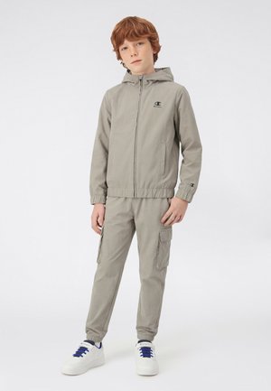 Ragazzo con capelli rossi che indossa una giacca con cappuccio grigio chiaro con zip e pantaloni cargo abbinati, in piedi con le mani rilassate, sneaker bianche con lacci blu.