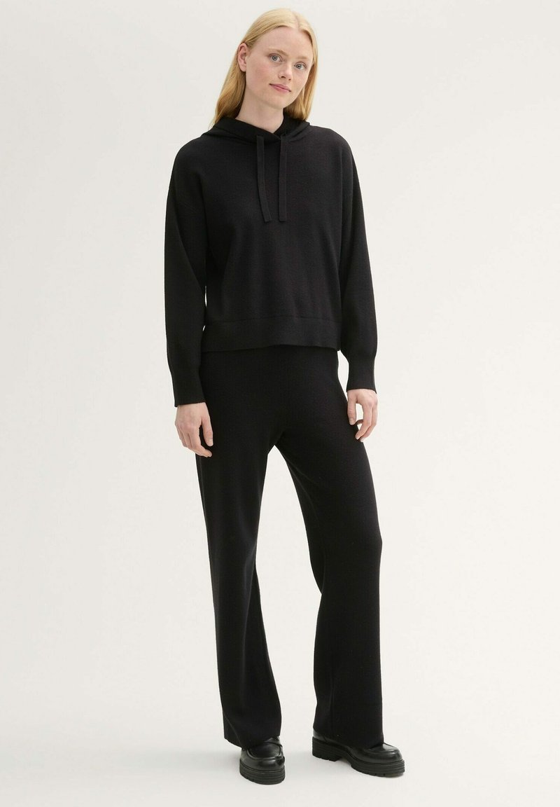 Schwarzer gestrickter Hoodie mit einer Fronttasche und Kordelzug, kombiniert mit schwarzen, figurbetonten Hosen. Beide Kleidungsstücke haben eine glatte Textur.