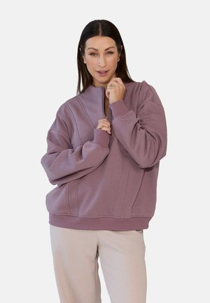 Kobieta z długimi ciemnymi włosami, ubrana w wrzosowy sweter z półgolfem, trzymająca zamek pod brodą, zestawiony z jasnobeżowymi spodniami.