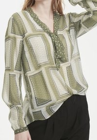 Zelená vzorovaná blůza s dlouhými rukávy, se vorovým výstřihem a mašlí u límce. Materiál je lehký a poloprůsvitný s puntíky.