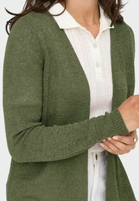 Grüne Strickjacke mit weicher Textur, offener Front und gerippten Bündchen, getragen über einem hellen Hemd mit Kragen.