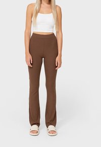 Pantalons en ribbed marron avec un bas évasé, associés à un crop top blanc et des sandales à glissière beiges. Le matériau semble doux et texturé.