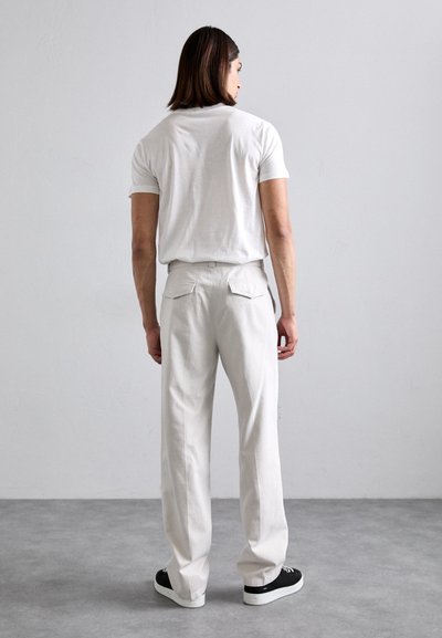Man with shoulder-length hair wearing white t-shirt, beige pants, and black sneakers standing facing a plain light gray wall.