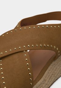 Καφέ σουέντ σανδάλι με λουριά που διασταυρώνονται, χρυσά καρφιά που τονίζουν τις άκρες και πλεκτή σόλα εσπαντρίγια.