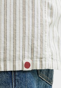 Gestreepte beige en witte zoom van een shirt met een klein stoffen label met een effen rode cirkel, gedragen over een blauwe spijkerbroek.