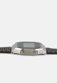 Montre numérique avec un écran rectangulaire noir, un boîtier en métal argenté et un bracelet en caoutchouc noir texturé. Dispose de deux boutons latéraux pour les réglages.