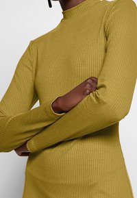 Rippstrick-Turtleneck-Top in Olivgrün, mit langen Ärmeln und einer figurbetonten Silhouette. Der Stoff hat eine glatte, strukturierte Oberfläche.