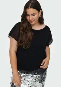 Sort top med korte, gennemsigtige ærmer, rund halsudskæring og blød struktur, parret med en sølvfarvet palietskørt for et kontrastfyldt look.