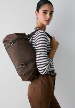 Mujer con el cabello peinado hacia atrás, lleva un suéter a rayas y pantalones marrones, y lleva una mochila grande de cuero marrón oscuro sobre un hombro.